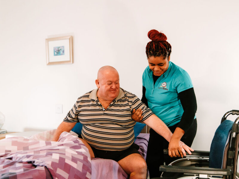 Smiling nurse assisting senior man to get up from bed. Caring nurse supporting patient while getting up from bed and move towards wheelchair at home. Helping elderly disabled man standing up in his bedroom.
